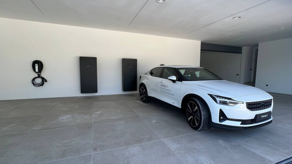 Un coche el&eacute;ctrico aparcado c&oacute;modamente en un garaje con una estaci&oacute;n de carga para veh&iacute;culos el&eacute;ctricos instalada en la pared.  Dos bater&iacute;as solares junto al cargador sugieren un sistema energ&eacute;tico dom&eacute;stico sostenible listo para alimentar el veh&iacute;culo el&eacute;ctrico.