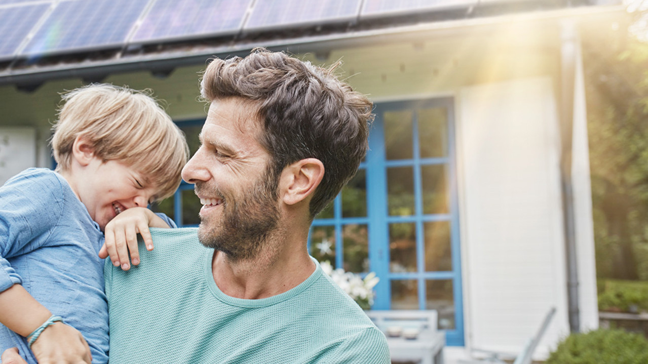 A man standing outside in the garden with a child on his arm, in the back we see a house with PV panels on the roof